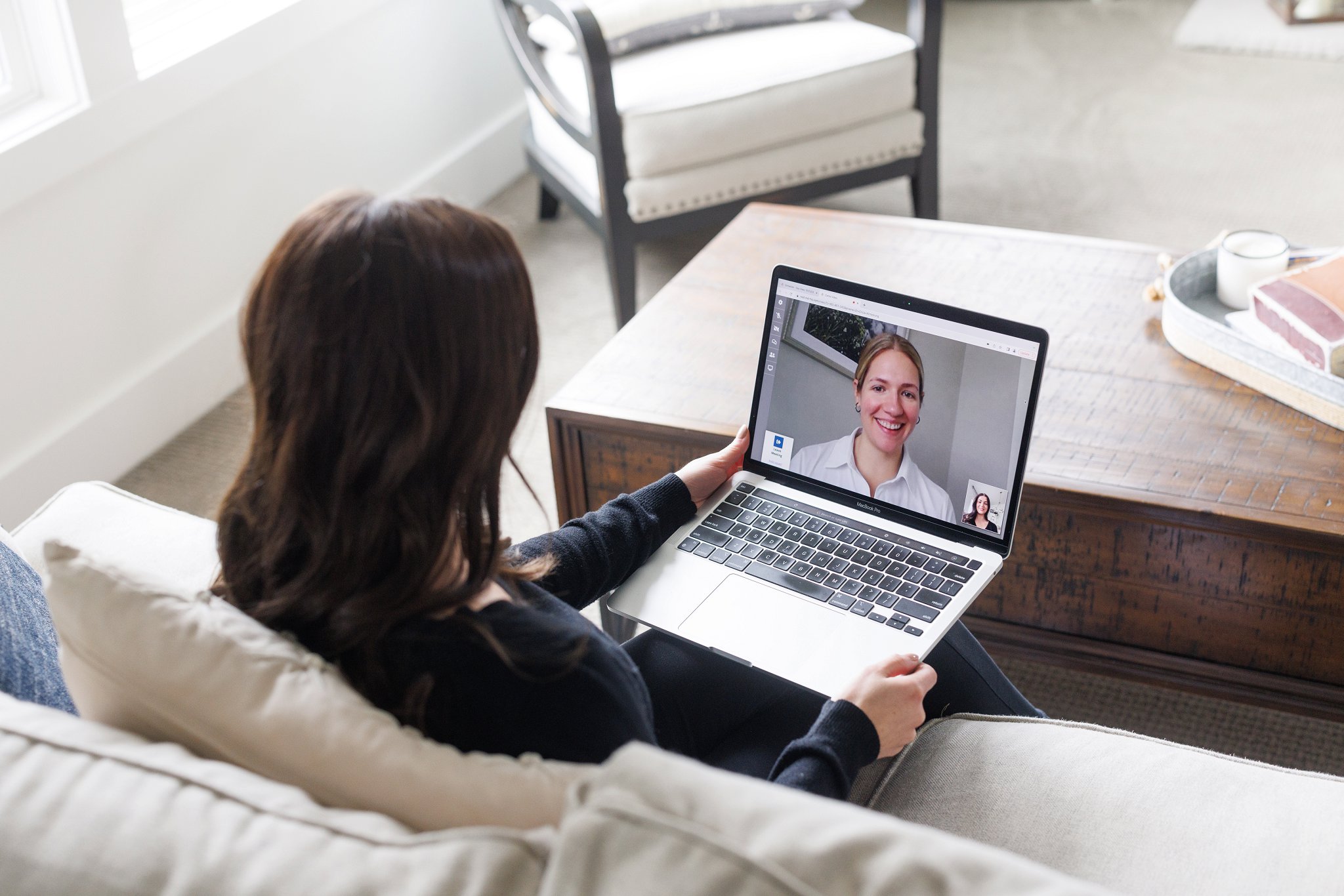 woman sitting on the couch, back to camera, looking at laptop screen, on the screen blonde woman smiling